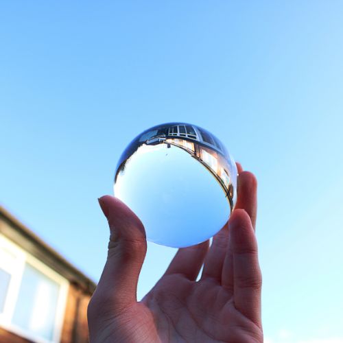 Eine Hand hält eine Glaskugel vor blauem Himmel. Ein Gebäude spiegelt sich darin.